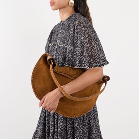 Model carrying a tan suede half-moon shoulder bag with whipstitch trim and knotted strap, styled with a dark floral dress