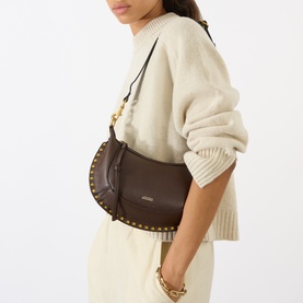 Model carrying Isabel Marant Oskan Moon Shoulder Bag in coffee leather with gold stud trim and adjustable strap, styled with cream knit and wide-leg