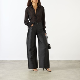 Model wearing Marques Almeida Mikado Boyfriend Trousers in black satin with wide-cut legs, styled with a sheer black shirt and heeled sandals.