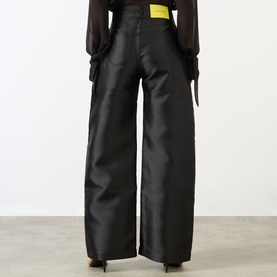Model wearing Marques Almeida Mikado Boyfriend Trousers in black satin, wide-leg cut with cargo-style side pockets and yellow logo badge.