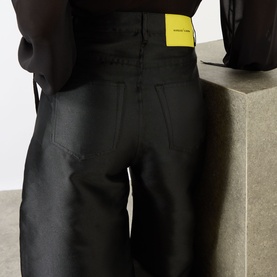 Model wearing Marques Almeida Mikado Boyfriend Trousers in black satin, rear view showing logo badge, patch pockets and wide-cut legs.