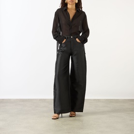 Model wearing Marques Almeida Mikado Boyfriend Trousers in black satin with wide-cut legs, styled with a sheer black shirt and heeled sandals.