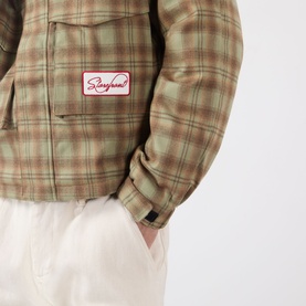 Model wearing Storefront Script Logo Flannel Utility Jacket in green check, close-up of logo patch and flap pocket detail, styled with white trousers.