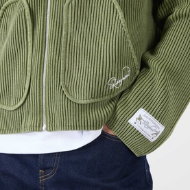 Model wearing Storefront Fisherman Script Logo Zip Sweater in olive ribbed knit, styled with white tee and navy jeans. Script logo and badge detail