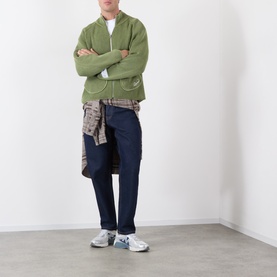 Model wearing Storefront Fisherman Script Logo Zip Sweater in olive, styled with navy trousers, plaid shirt tied at waist and white trainers.