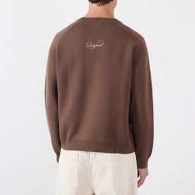 Model wearing Storefront brown cotton knit sweater, rear view showing embroidered logo at upper back, styled with cream trousers
