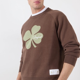 Model wearing Storefront brown cotton sweatshirt with sage green four-leaf clover graphic print, layered over white crew neck tee