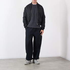 Model wearing Storefront Fisherman Script Logo Sweater in Charcoal, styled with black bomber jacket, wide-leg trousers and grey trainers.