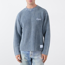 Model wearing Storefront Fisherman Script Logo Sweater in blue, ribbed knit, crew neck, with distressed denim jeans.