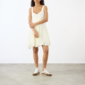 Model wearing JW Anderson ivory sleeveless mini dress with pleated skirt and sweetheart neckline, styled with white trainers.