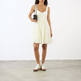 Model wearing JW Anderson ivory mini dress with sweetheart neckline and full skirt, styled with a dark shoulder bag and white trainers.