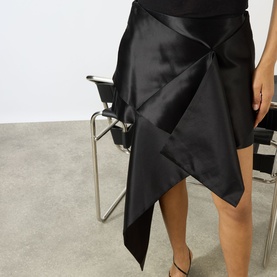 Model wearing JW Anderson Cross Drape Asymmetrical Mini Skirt in black, with origami-style draped front and asymmetric hemline in silk-cotton satin.