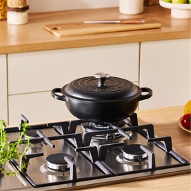 Le Creuset Matte Black cast iron casserole dish with silver knob lid, styled on a stainless steel gas hob in a kitchen setting.