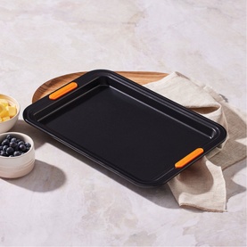 Le Creuset Small Baking Tray 27cm in Satin Black, rectangular non-stick carbon steel with orange silicone handles, styled on marble surface