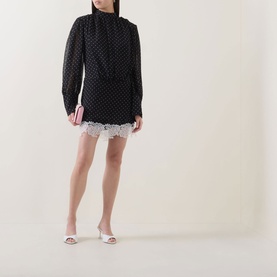 Model wearing Rotate black polka dot mini dress with lace hem trim, long sleeves and high neck, styled with white heeled mules