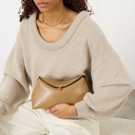 Model carrying Toteme T-Lock Leather Clutch in camel smooth leather with gold-tone T-lock fastening, styled with beige knit sweater and white trousers