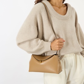 Model carrying Toteme T-Lock Leather Clutch in camel smooth leather with gold-tone T-lock fastening, worn crossbody with cream knit and trousers