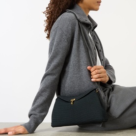 Model carrying Toteme T-Lock Rope Clutch in black with gold-tone T-lock fastening and woven finish, worn crossbody over grey knitwear