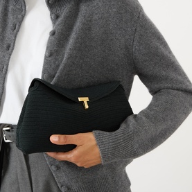 Model carrying Toteme T-Lock Rope Clutch in black with gold-tone T-lock fastening and woven finish, styled with grey cardigan