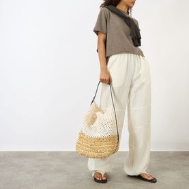 Model carrying Toteme Belted Raffia Tote Bag in Ecru/Vanilla, dual-toned raffia with leather belt strap, styled with grey tee and white wide-leg
