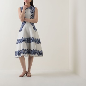 Model wearing Ulla Johnson dress in Lapis, white fit-and-flare silhouette with bold blue floral border print, styled with tan heeled sandals