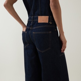 Model wearing Ulla Johnson Tigris jumpsuit, rear view showing indigo denim wide-leg style with tan leather logo patch pocket detail