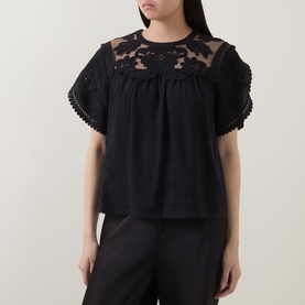 Model wearing Ulla Johnson Alma Embroidery Lace Silk Top in Noir, featuring floral lace yoke, short sleeves, and loose fit, styled with black trousers