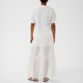 Model wearing Kaffe chalk white lace-panelled tiered maxi skirt, styled with a white short-sleeve T-shirt and flat sandals, rear view.