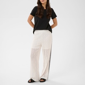 Model wearing Kaffe chalk wide-leg sheer lace trousers with black side stripe, styled with a black T-shirt and black sandals.