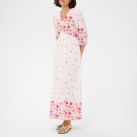 Model wearing Kaffe White Rose Border Flower maxi dress with puff sleeves, V-neckline, and pink floral border print, styled with tan sandals.