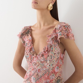 Model wearing Rixo dress in Butterfly Paisley Orange, 100% silk with V-neck, ruffle cap sleeves, styled with gold hoop earrings.