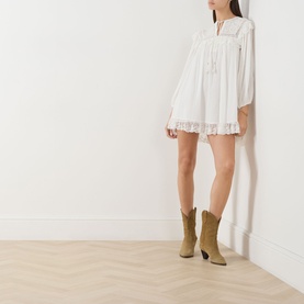 Model wearing Rixo ivory mini dress with lace trim, long sleeves and tie neckline, styled with tan cowboy boots