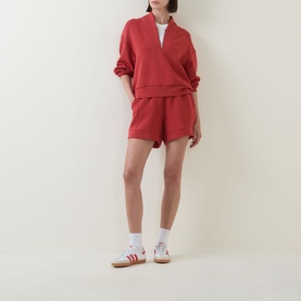 Model wearing red half-zip sweatshirt and matching shorts set, styled with white socks and Adidas trainers
