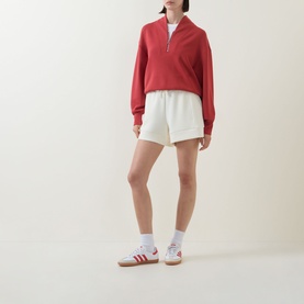 Model wearing Varley Pristine shorts in off-white, styled with a red half-zip sweatshirt and white socks with trainers.