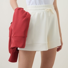 Model wearing Varley Pristine shorts in ivory with elasticated drawstring waist, styled with a red hoodie draped over arm
