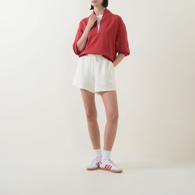 Model wearing Varley Pristine shorts with matching red half-zip sweatshirt and white Adidas trainers with red stripes.