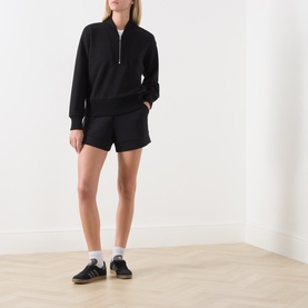 Model wearing Varley black shorts with matching black half-zip sweatshirt, white socks and navy trainers.