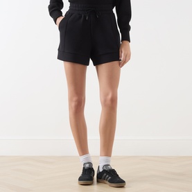Model wearing Varley black shorts with elasticated waist, styled with a black knit top, white socks and blue trainers.
