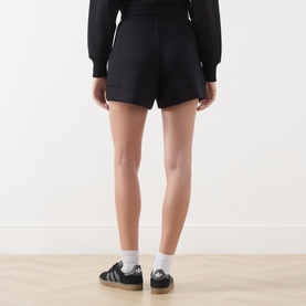 Model wearing Varley black shorts, rear view, styled with a black long-sleeve top, white socks and black Adidas Gazelle trainers.