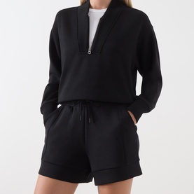 Model wearing Varley black shorts with drawstring waist, styled with matching black half-zip sweatshirt and white top.