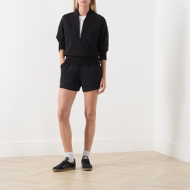 Model wearing Varley black shorts with matching black half-zip sweatshirt, white socks and black trainers.