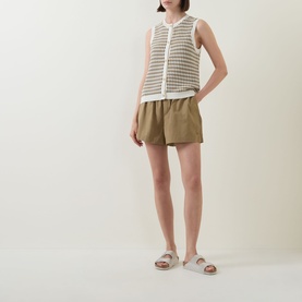 Model wearing Varley Golden Olive cotton shorts, styled with a white sleeveless open-knit top and grey flat sandals.