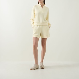 Model wearing Varley shorts in Anise Flower, a pale yellow hue, styled with a matching half-zip sweatshirt and cream trainers.