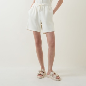 Model wearing Varley Pristine shorts in white, styled with chunky flatform sandals. Elasticated waistband with drawstring tie visible.