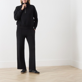 Model wearing Varley black half-zip sweatshirt and wide-leg trousers, styled with dark trainers on a light wood floor.