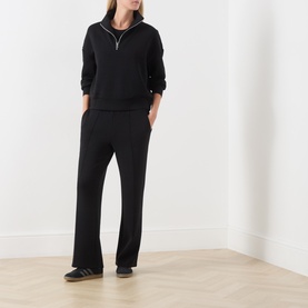 Model wearing Varley black half-zip sweatshirt with contrast trim, styled with matching wide-leg black trousers and grey trainers.