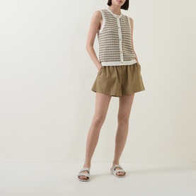 Model wearing Varley Aura Sweater Vest in Egret/Golden Olive check, styled with khaki shorts and white flat sandals.