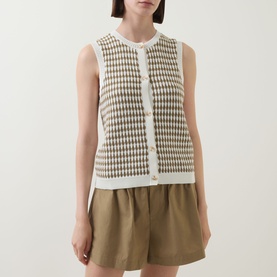 Model wearing Varley Aura Sweater Vest in Egret/Golden Olive check, sleeveless with button front and round neck, styled with khaki shorts.