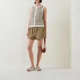 Model wearing Varley Aura Sweater Vest in Egret/Golden Olive check, styled with khaki shorts, slide sandals and small crossbody bag.