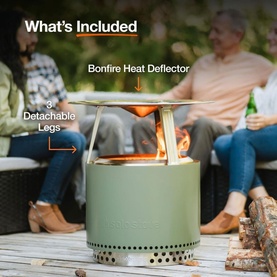 Solo Stove Bonfire Heat Deflector shown fitted to a green fire pit with 3 detachable legs, flames visible inside, outdoors lifestyle setting.